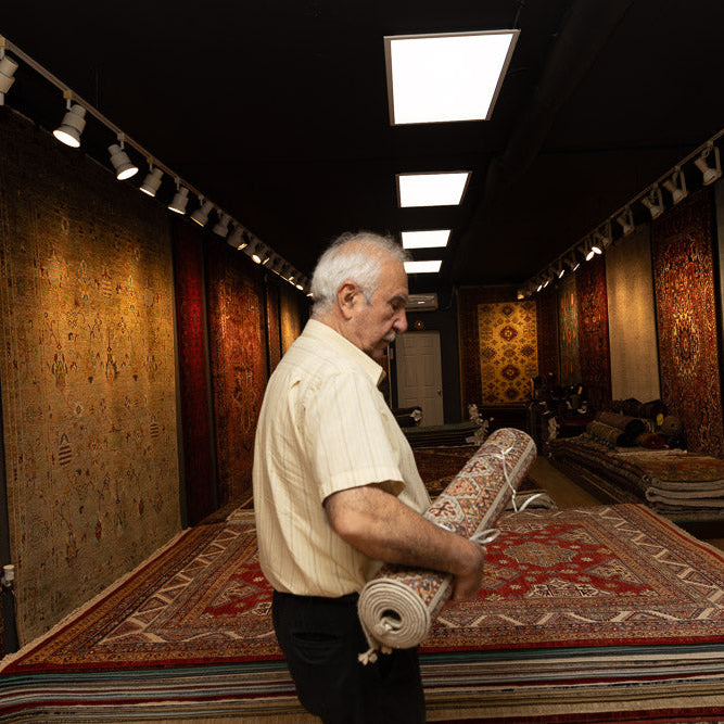 Man holding a rolled-up rug in a room with various rugs displayed on the walls and floor.